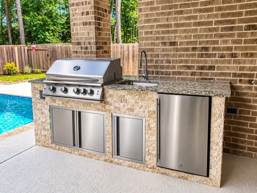 Outdoor kitchen with stainless grill, sink and refrigerator beside a pool